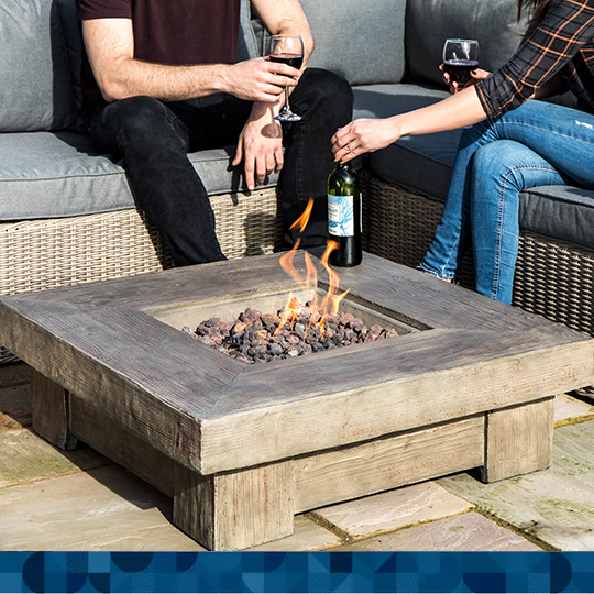 A square shaped gas fire pit sits low to a slate surface with two people sitting on furnishings next to it.