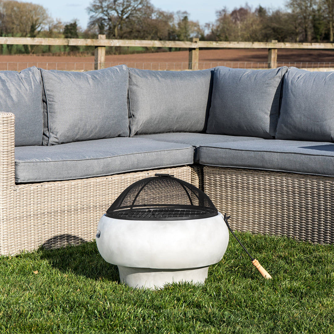 A stone look gas fire pit with flames burning brightly in a garden with a white cup on the fire pit table.