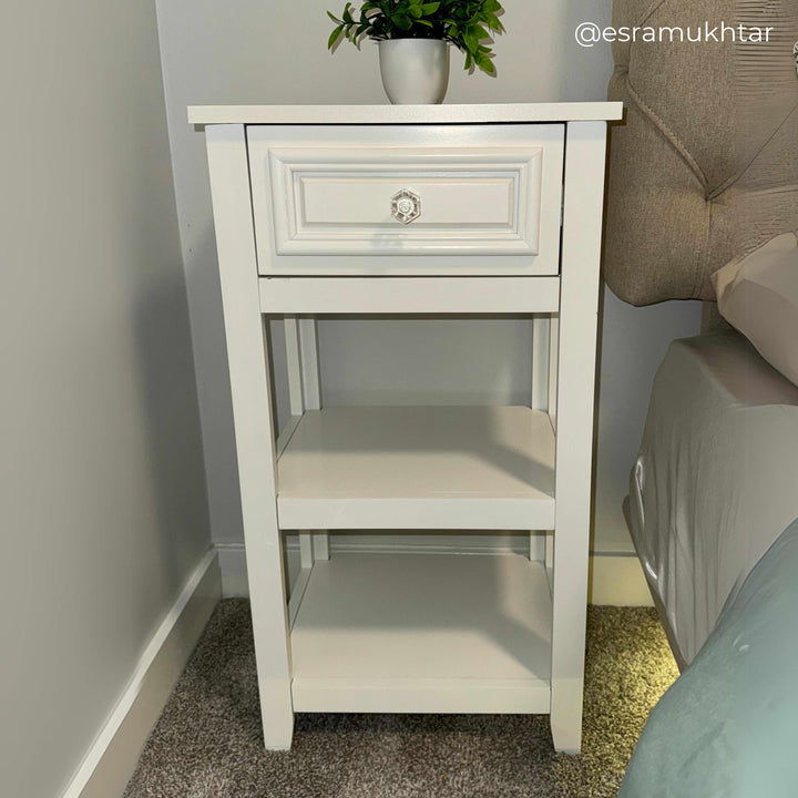 White nightstand with a drawer and two shelves next to a bed