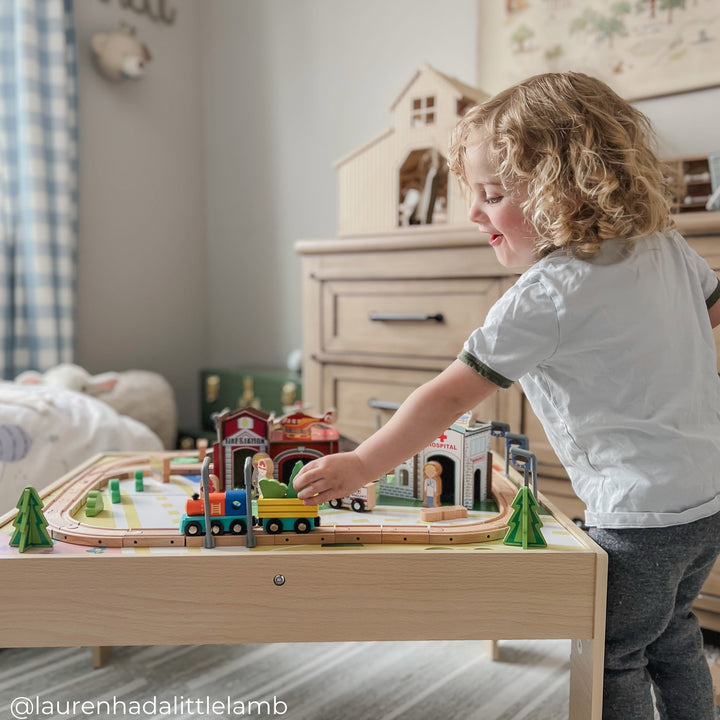 A young child with curly hair plays with a wooden train set on a table in a neatly decorated room. A bed with a stuffed animal is visible in the background.