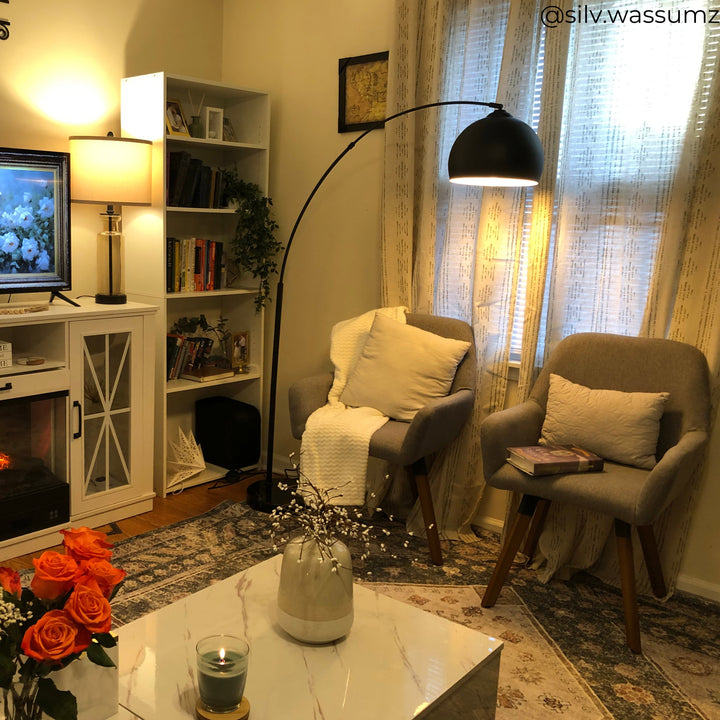 A cozy living room with two armchairs, a floor lamp, a small bookcase, and a TV. A vase of roses is on a marble coffee table. Light filters through sheer curtains.