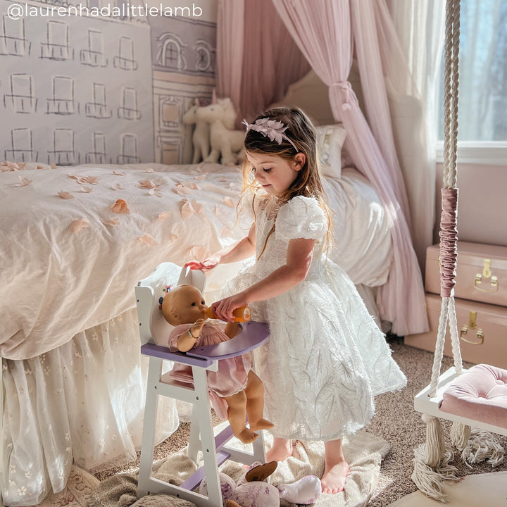 A little girl feeds her baby doll a bottle, sat inside a baby doll highchair