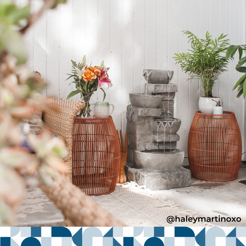 Teamson grey stone stacked fountain is on an outdoor patio surrounded by two wicker outdoor tables.