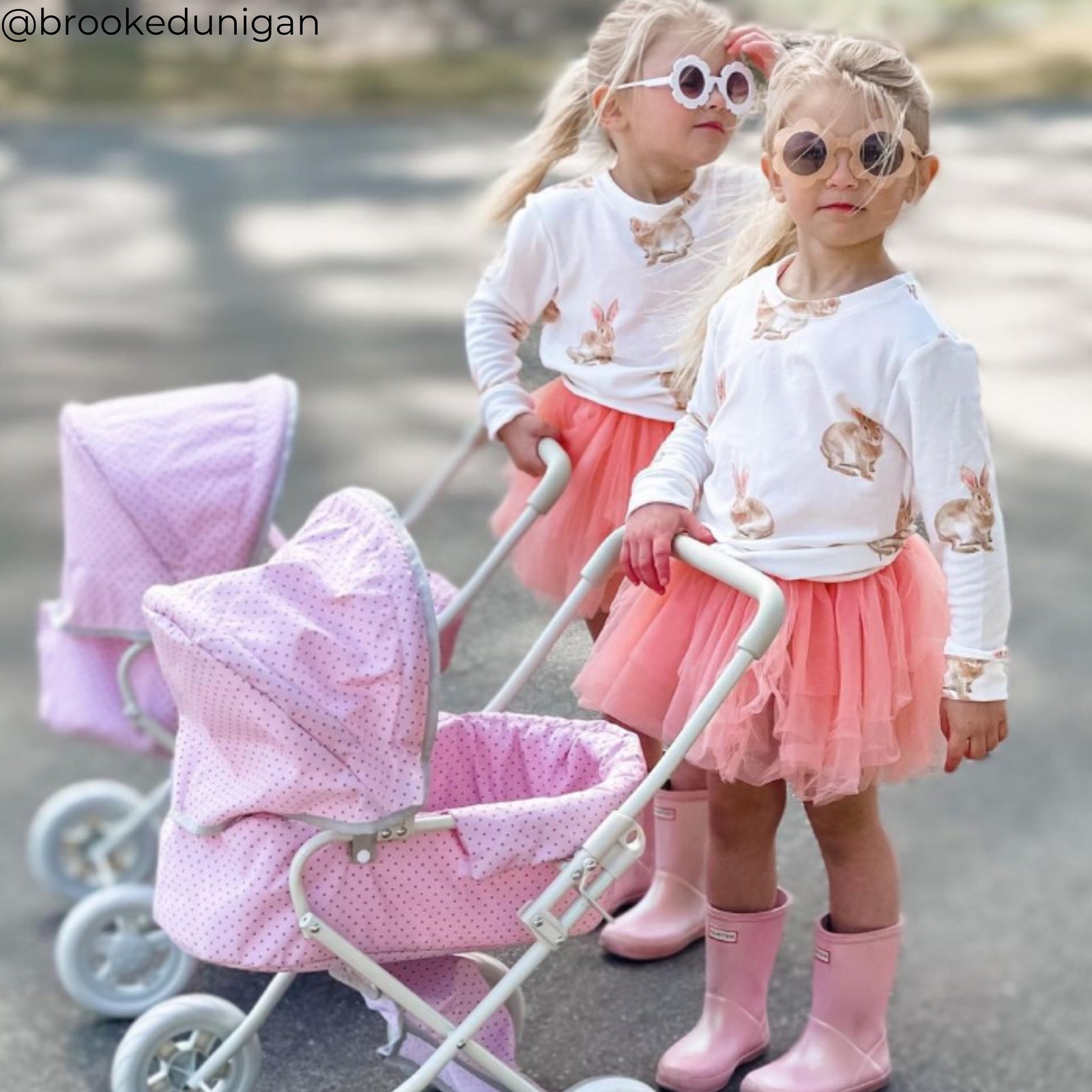 Two little girls in pink tutus and sunglasses pushing doll strollers on a street.