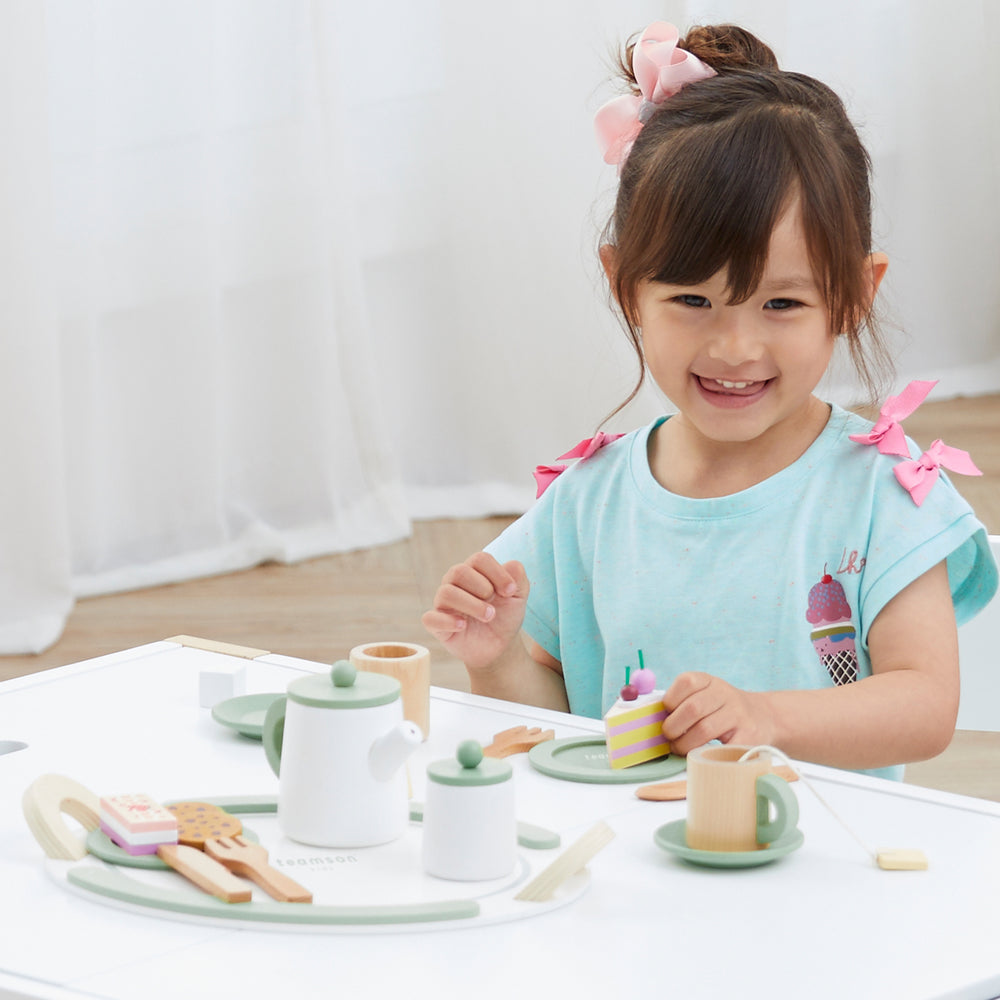 A young child with a ponytail sits at a table, smiling while playing with a toy tea set from Teamson Kids. The child wears a light blue shirt with pink bows on the shoulders. The room has a white, airy background.