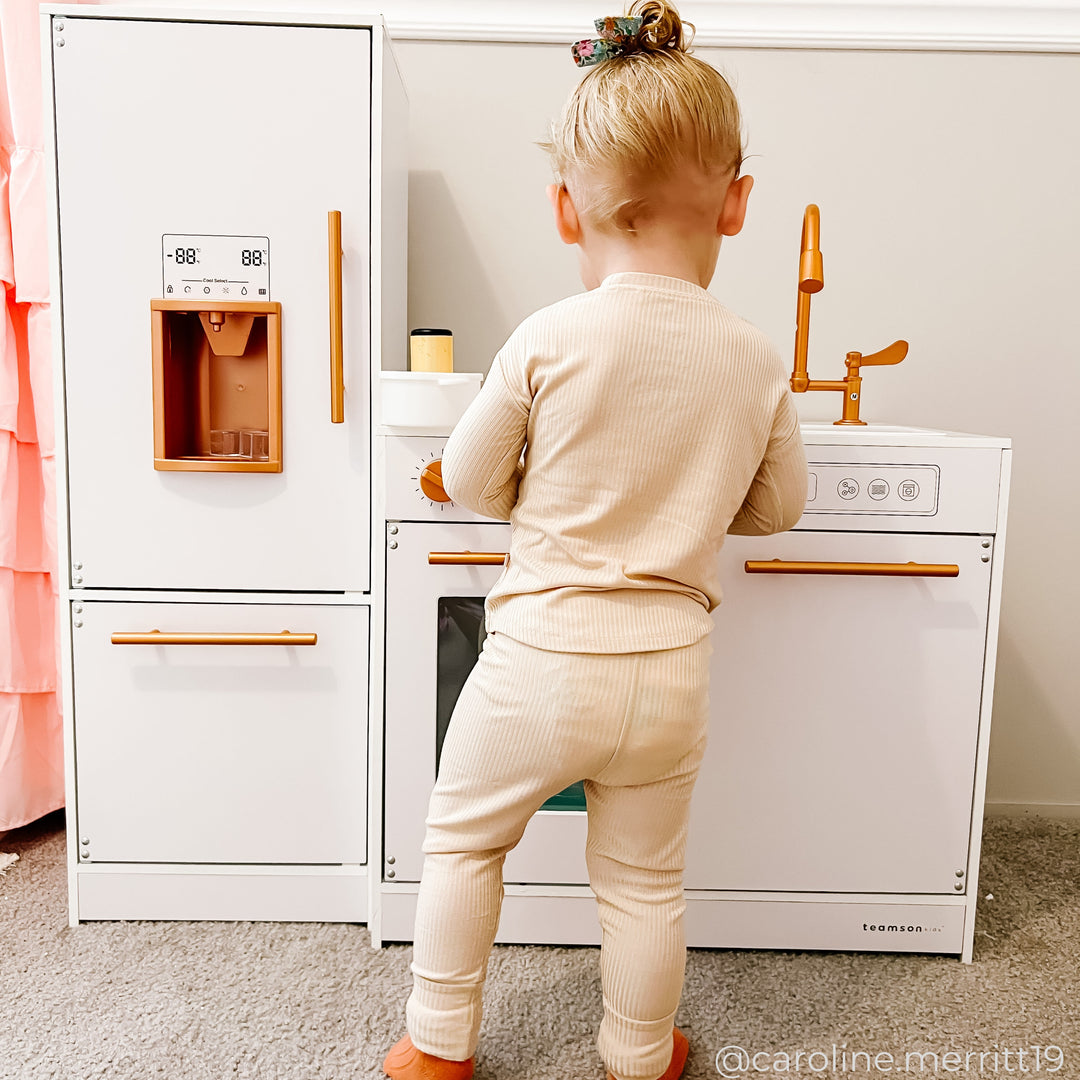 A young child dressed in beige pajamas stands facing a white play kitchen set with a fridge, stove, and sink, engaging with the setup. The child's hair is tied up in a small bun.