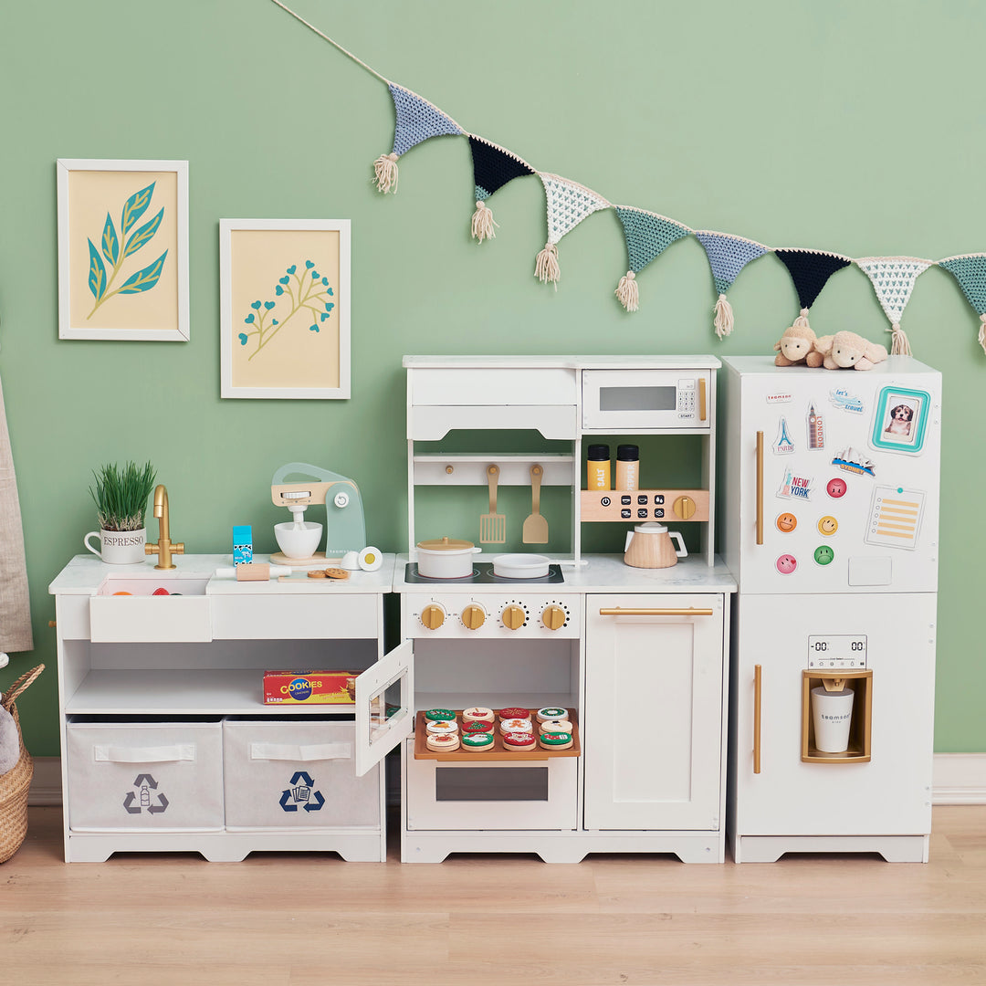 Sentence with Product Name: Teamson Kids - Little Chef Atlanta Large Modular Play Kitchen, white/gold arranged against a wall with accessories and decorations, creating a playful simulation of a kitchen environment for children.