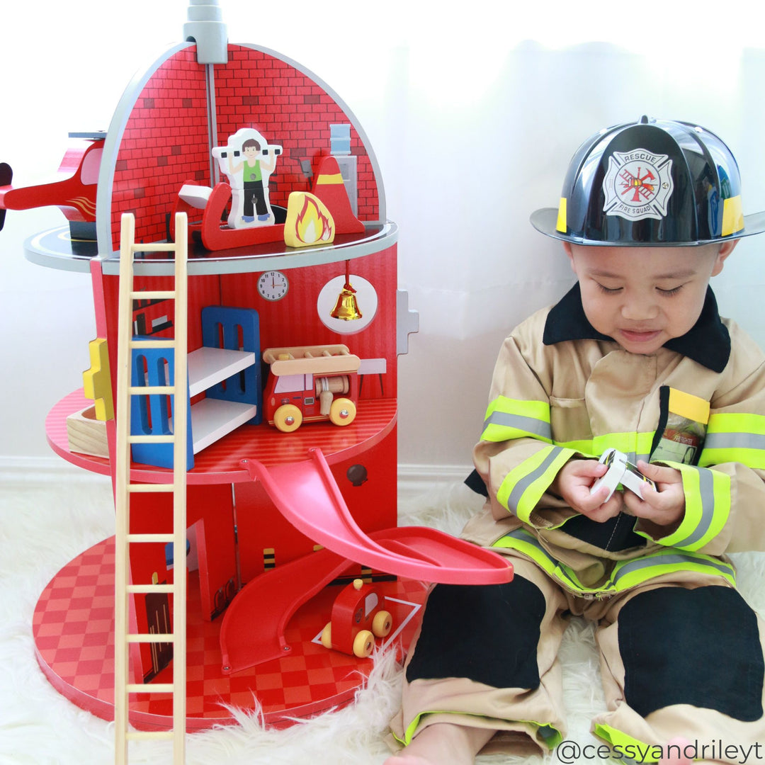 A child dressed in a firefighter costume is sitting down holding a firefighter figure next to a toy fire station playset with a slide, a ladder, and various accessories.