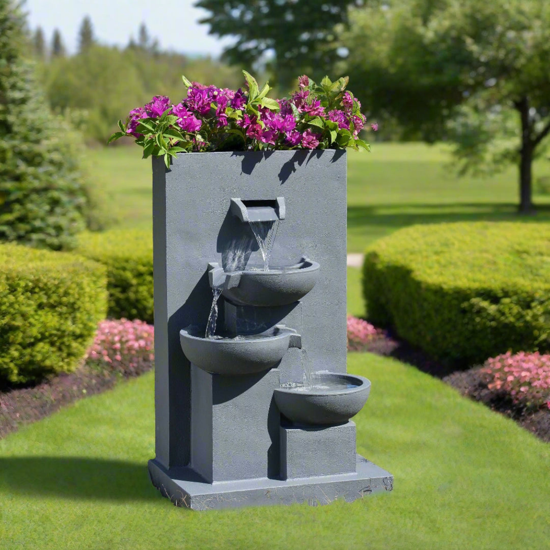 29.13" Outdoor Water Fountain with Planter & LED Lights, Matte Gray with purple flowers on top, set against a natural backdrop with a pond.