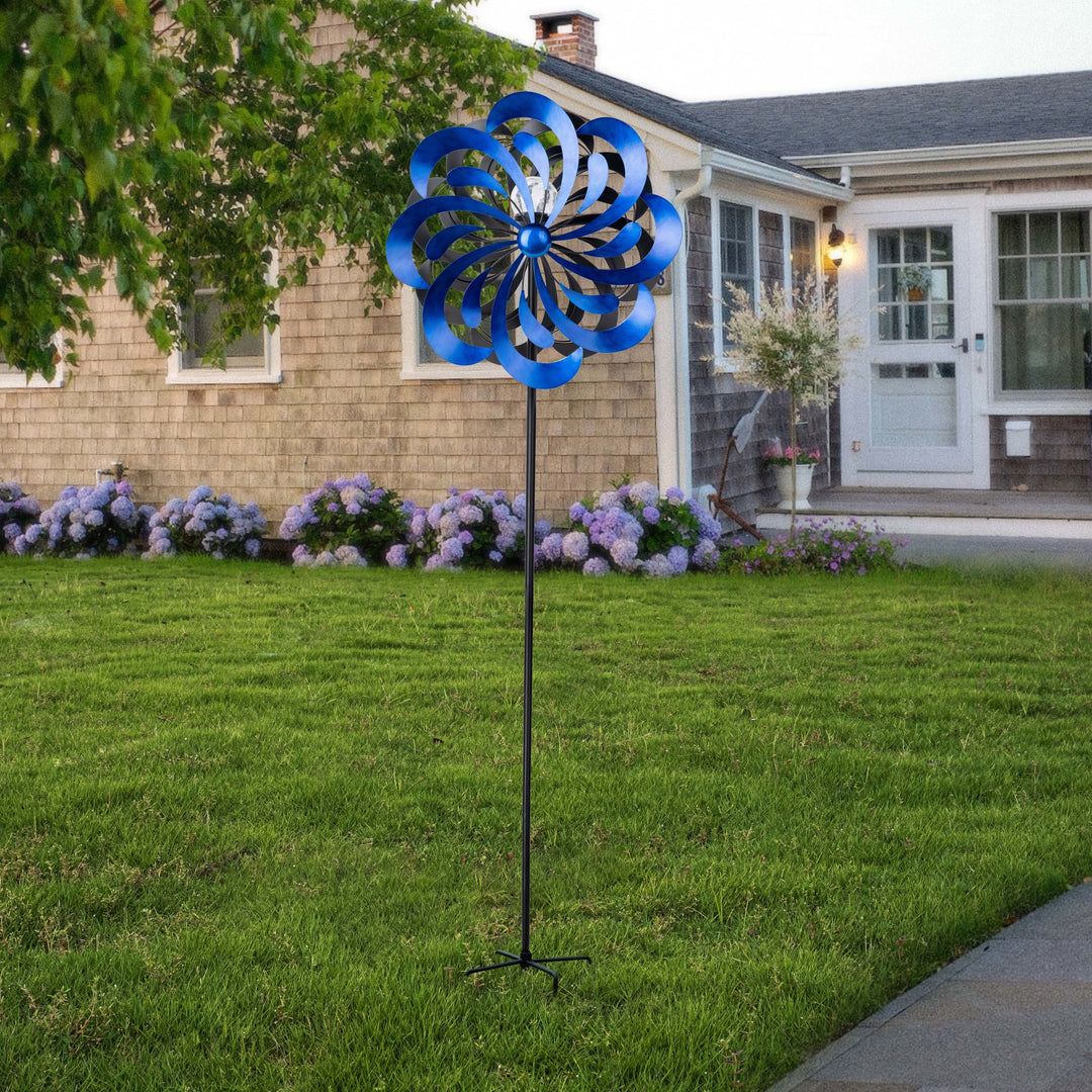 A Blue 18" dia. x 70" H Solar Metallic Kinetic Windmill Spinner stands in front of a house with hydrangea bushes.
