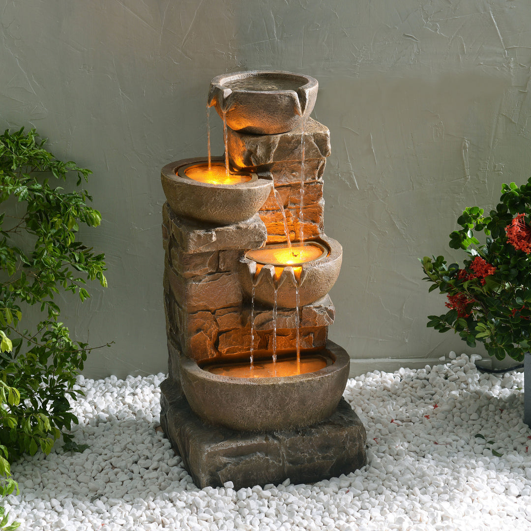 An illuminated 33.27" Cascading Bowls & Stacked Stones LED Outdoor Fountain with flowing water, nestled among plants and white pebbles.
