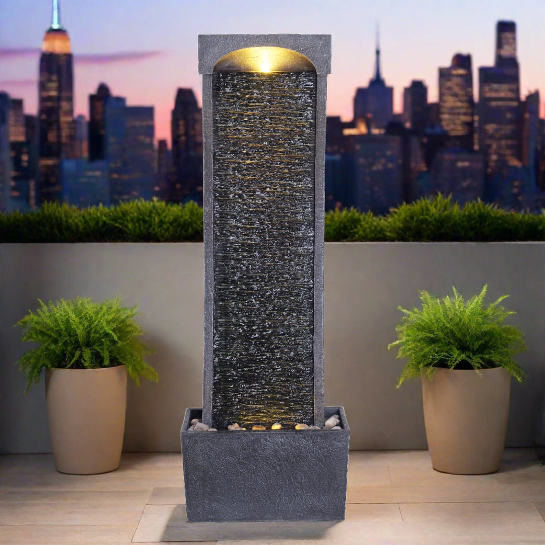 A waterfall fountain, charcoal, with LED lights illuminated at the top and the bottom, on a balcony, with a skyline behind it.
