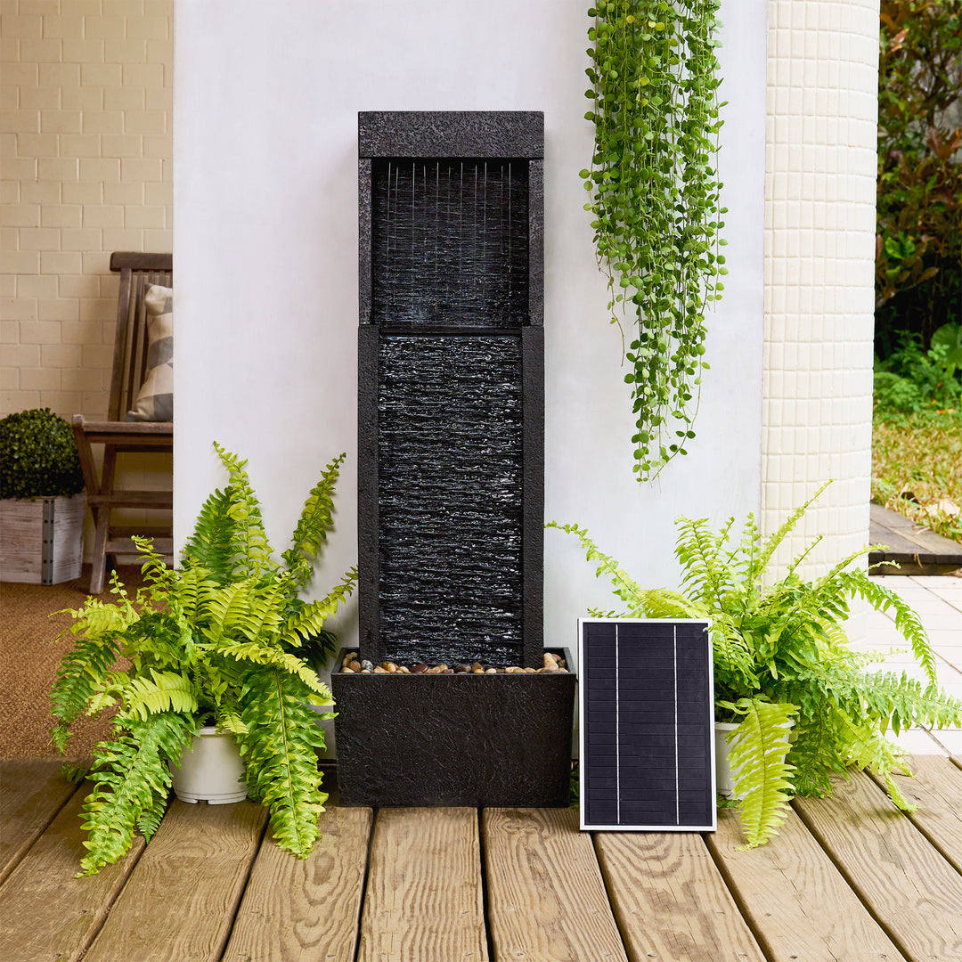A vertical water feature with dark slate finish is placed on a wooden deck, surrounded by ferns and a hanging plant. A small solar panel rests against the fountain's base.
