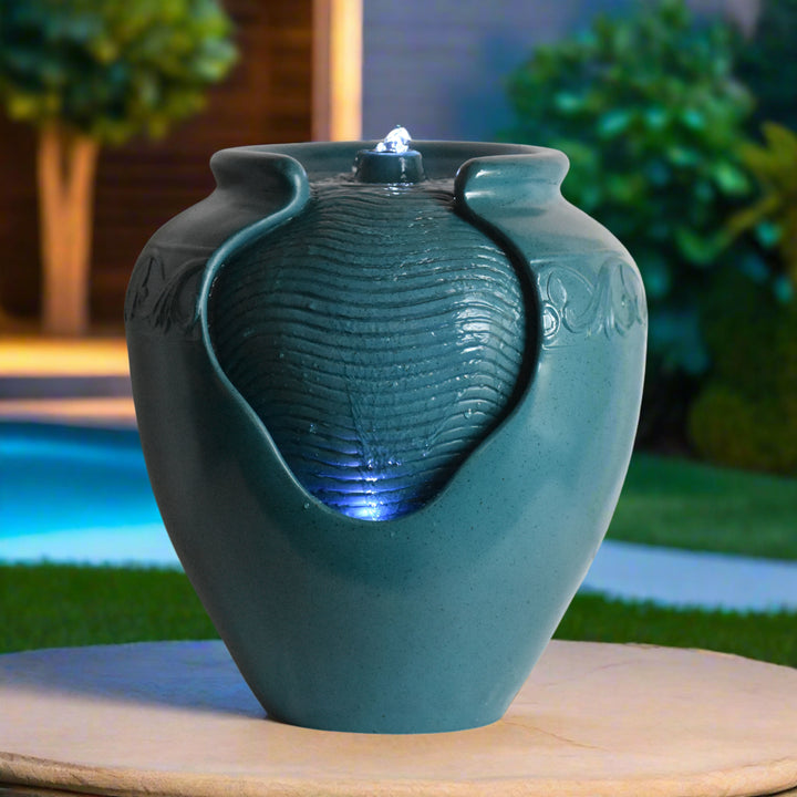 A close-up of a teal vase-shaped water fountain at night with LED lighting illuminating the textured surface