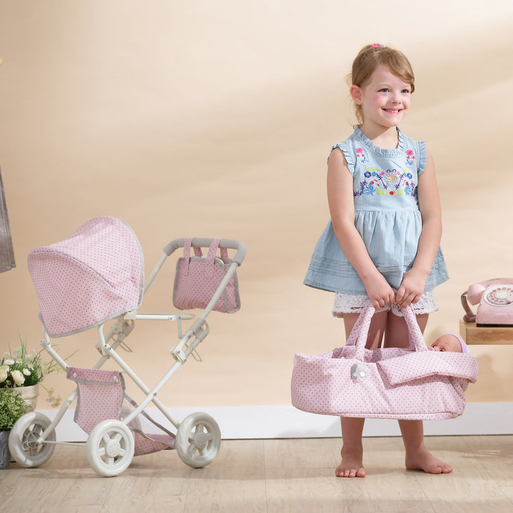 A little girl in a blue shirt and white shorts is holding the removable bassinet in her hands and she is standing next to the frame of the baby doll buggy that has the canopy, storage basket and diaper bag still attached.