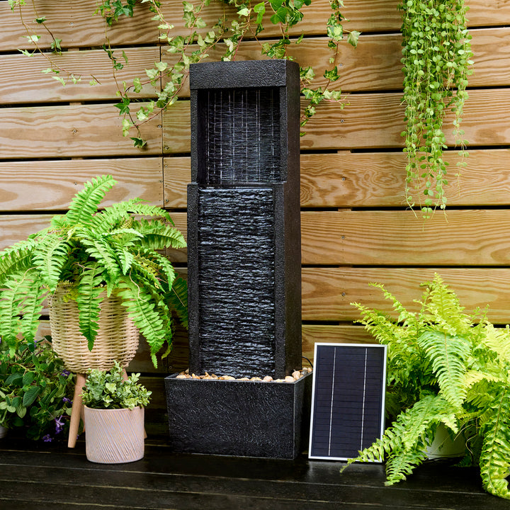 A Teamson Home Athena Waterfall Fountain, textured black and a base with pebbles, surrounded by potted plants