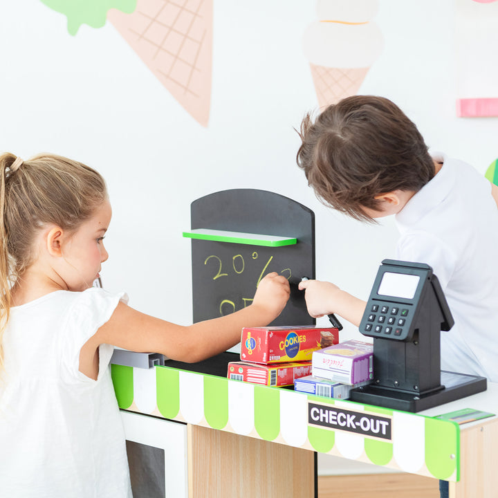 Two kids playing store at a pretend market stand with groceries and a Teamson Kids Cashier Austin Play Market Checkout Counter with 26 Accessories, Green/Natural.