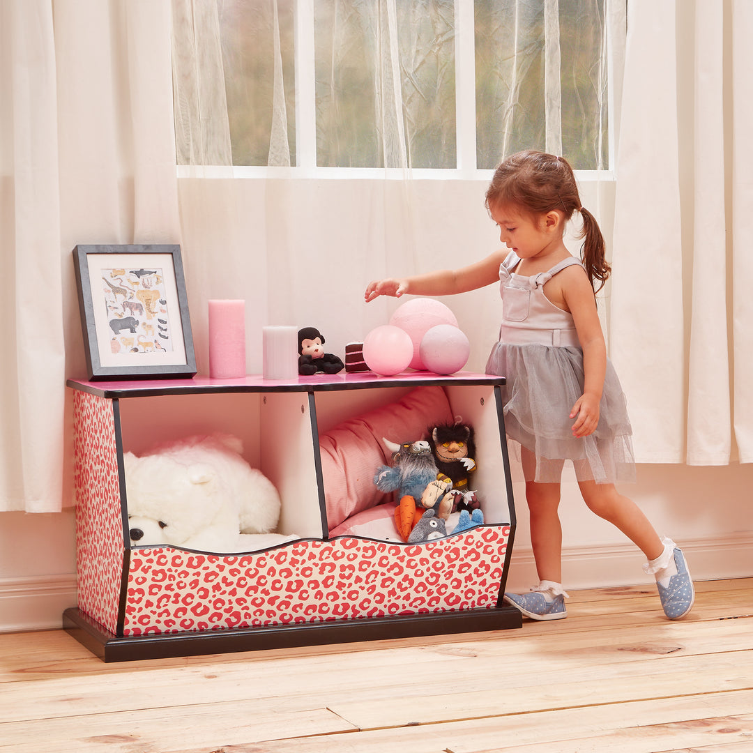 A little girl with a white top and gray tutu stands next to a pink and black two-cubbies storage bench with pink leopard print accents. filled with stuffed animals.