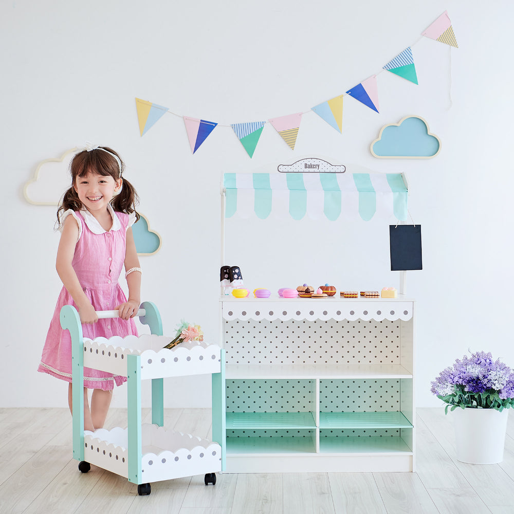 A girl stands behind the rolling dessert cart which has flowers on top, and next to a bakery stand of treats in a children's playroom.