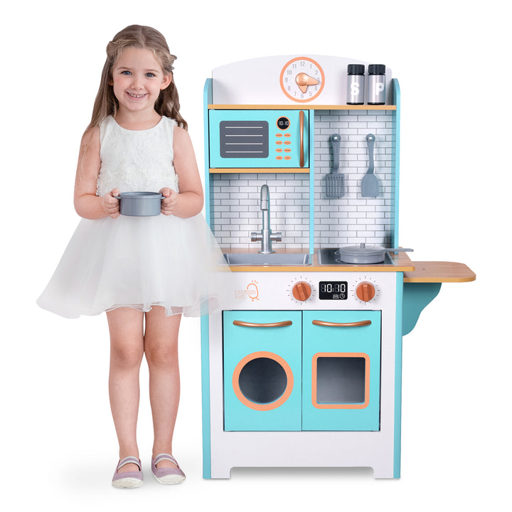 A smiling young girl standing next to a Teamson Kids Little Chef Santos Retro Wooden Kitchen Playset in Aqua/White with interactive features.