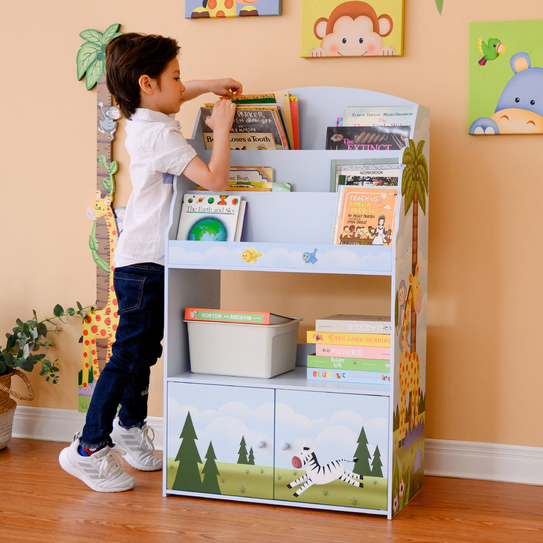 A boy standing in front of a Fantasy Fields Sunny Safari Kids 3-Tier Wooden Bookshelf with Storage, Multicolor.