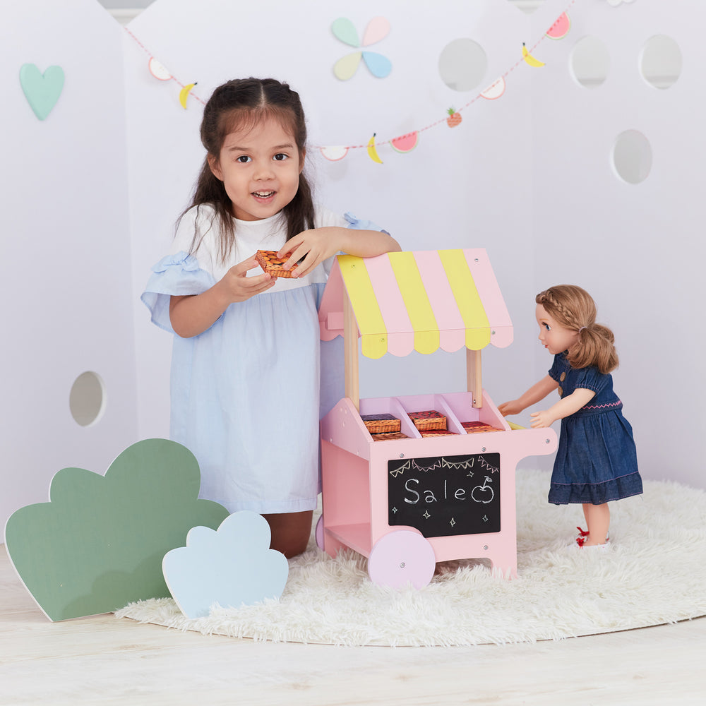 A little girl in blue and white kneeling next to an 18" doll fruit stand, pink and yellow with a chalkboard, and a doll in a blue dress.