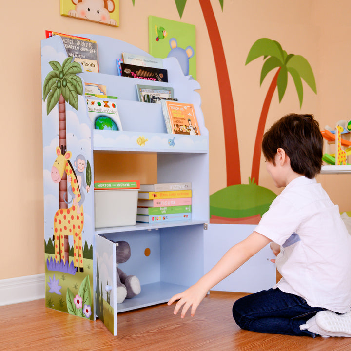 The boy is sitting on the floor near the Fantasy Fields Sunny Safari Kids 3-Tier Wooden Bookshelf with Storage, Multicolor.