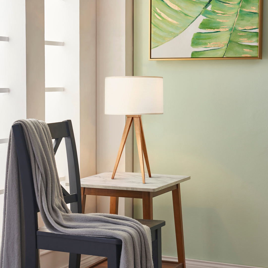 A Teamson Home Romanza 20" Natural Wood Tripod Table Lamp with White Drum Shade on a end table next to a gray chair in a green room with a big window behind it