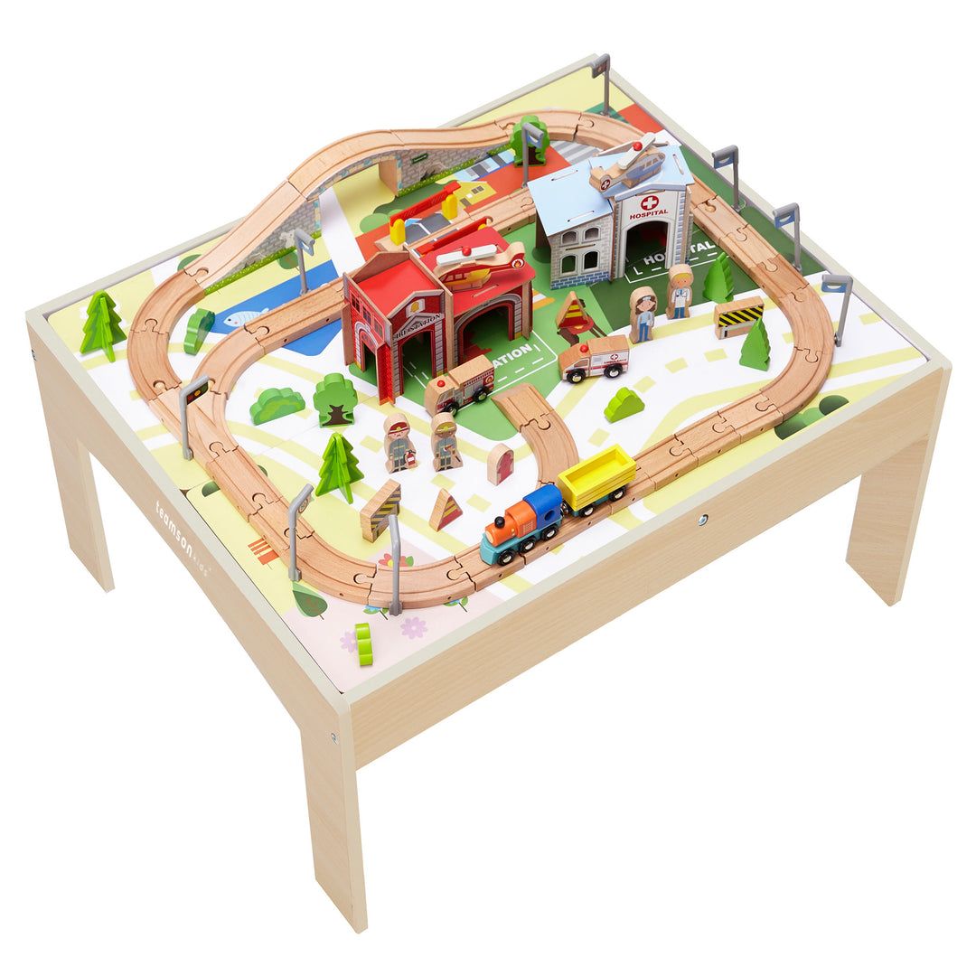 Teamson Kids Preschool Play Lab Toys Wooden Table with 85-pc Train and Town Set, Natural on a table with track arrangement, a station, trees, and toy figures from above.