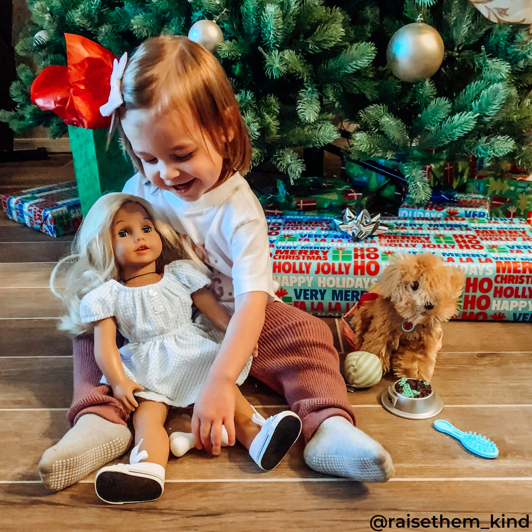 A little girl next to a Christmas tree holding a new doll next to a plush puppy dog.