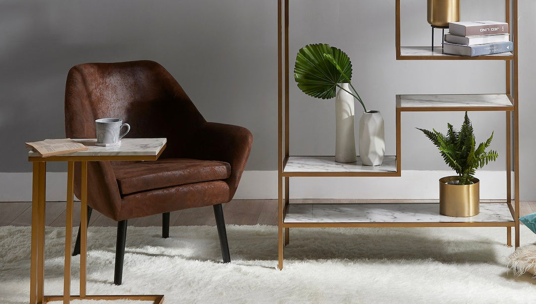 A brown leather accent chair in between a side table and a bookshelf with gold frame work and faux marble shelves.