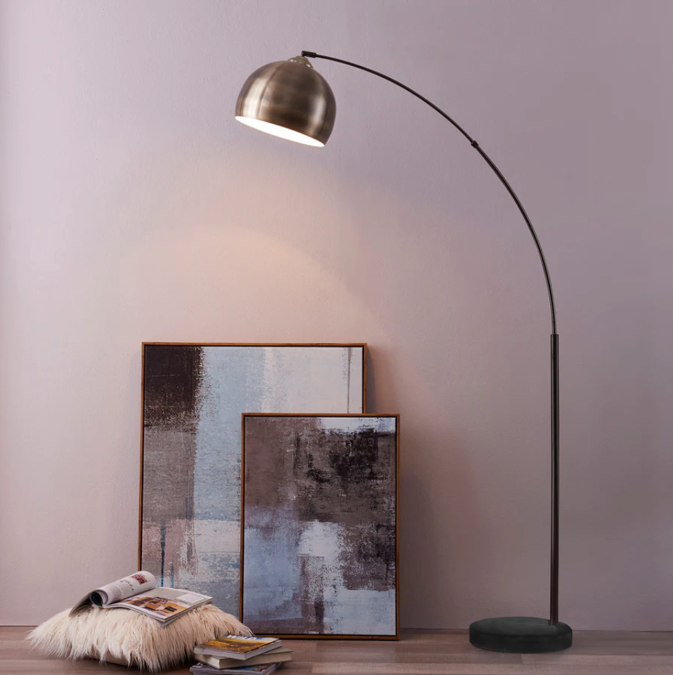Arc floor lamp with a brass shade shines on paintings leaning against a wall