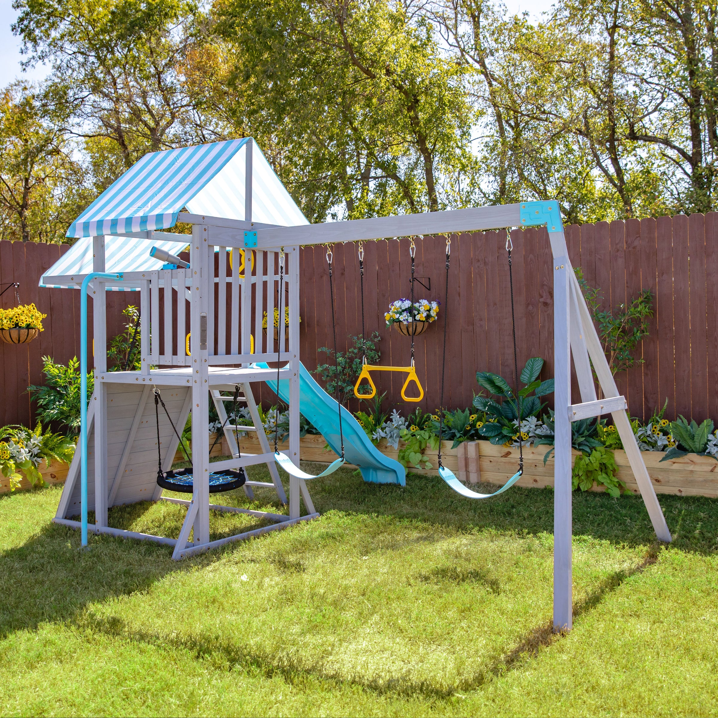 Playground set with swings, slide, and climbing area on grass. Surrounded by a wooden fence and plants in a backyard. Blue-striped canopy on top.