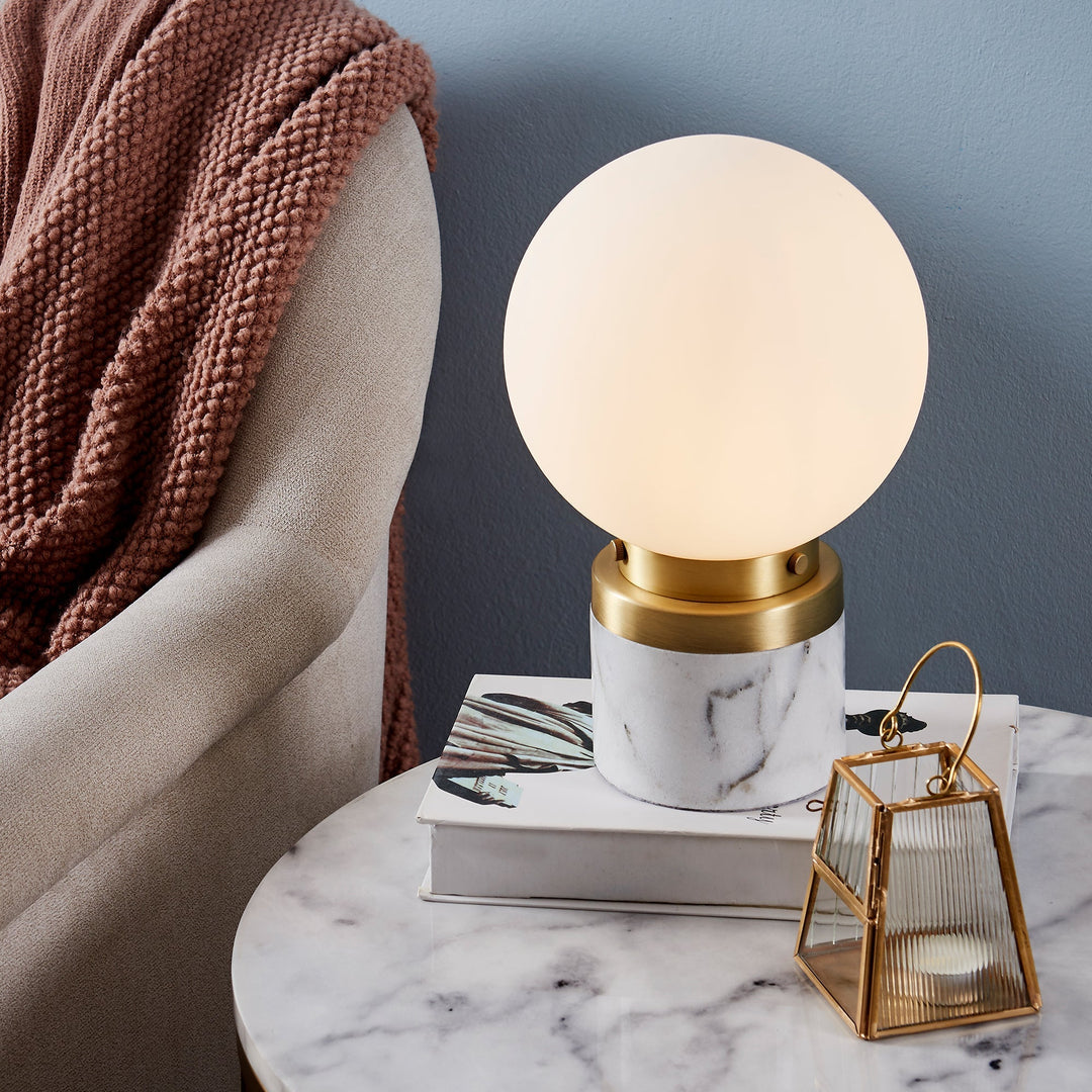 A globe-shaped table lamp with a faux marble and gold base sitting on top of a faux marble side table.