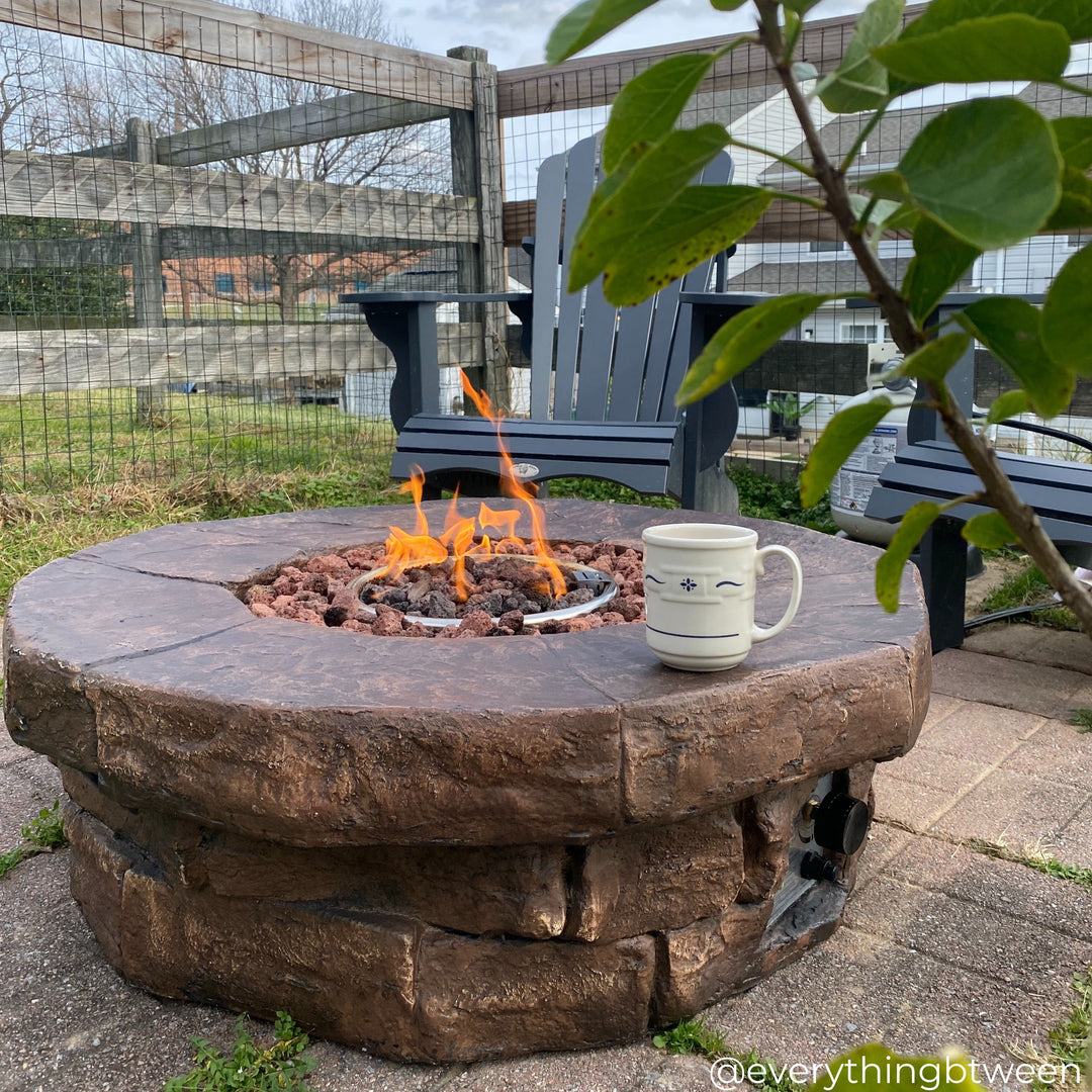 A stone look gas fire pit with flames burning brightly in a garden with a white cup on the fire pit table.