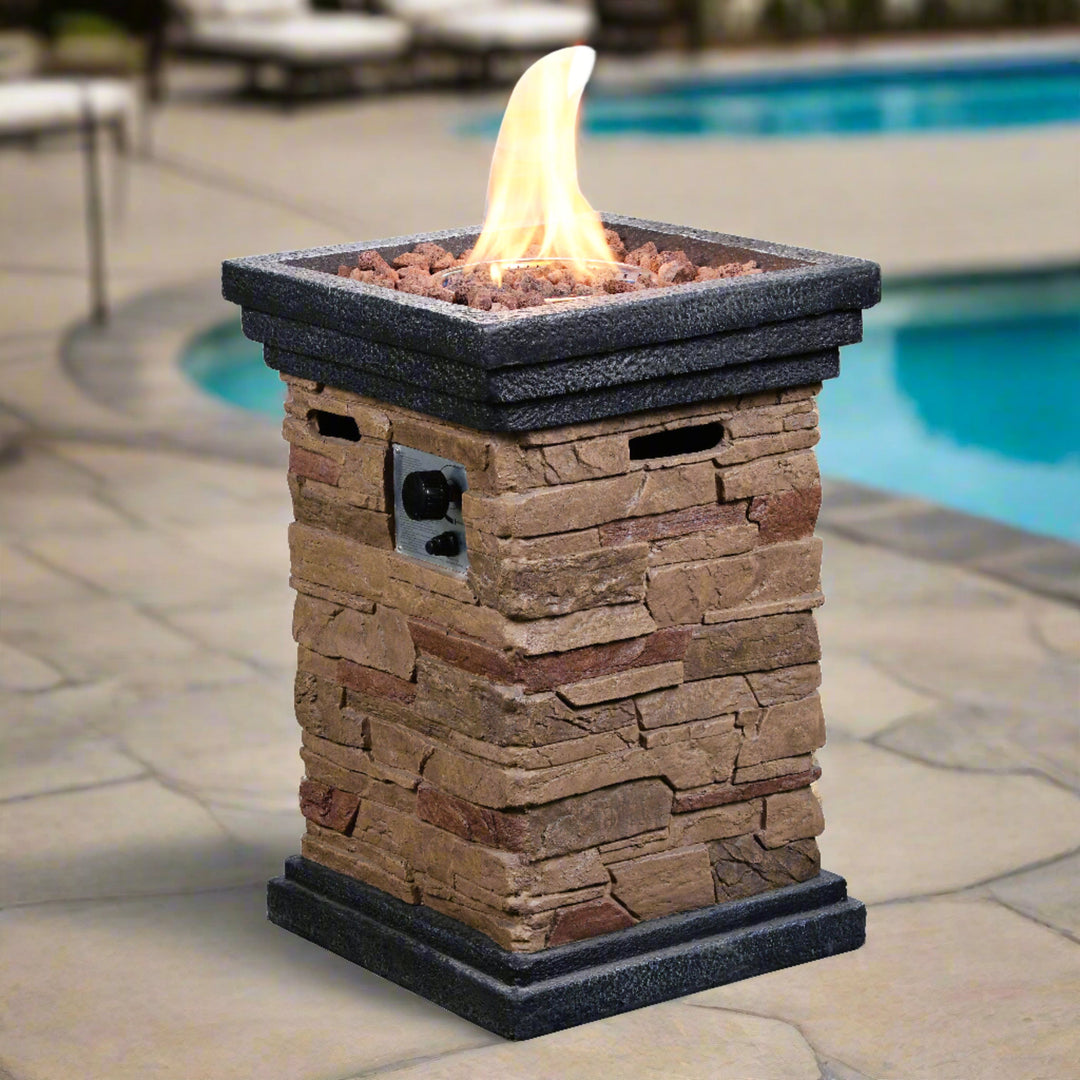A stone look gas fire pit with flames burning brightly in a garden with a white cup on the fire pit table.