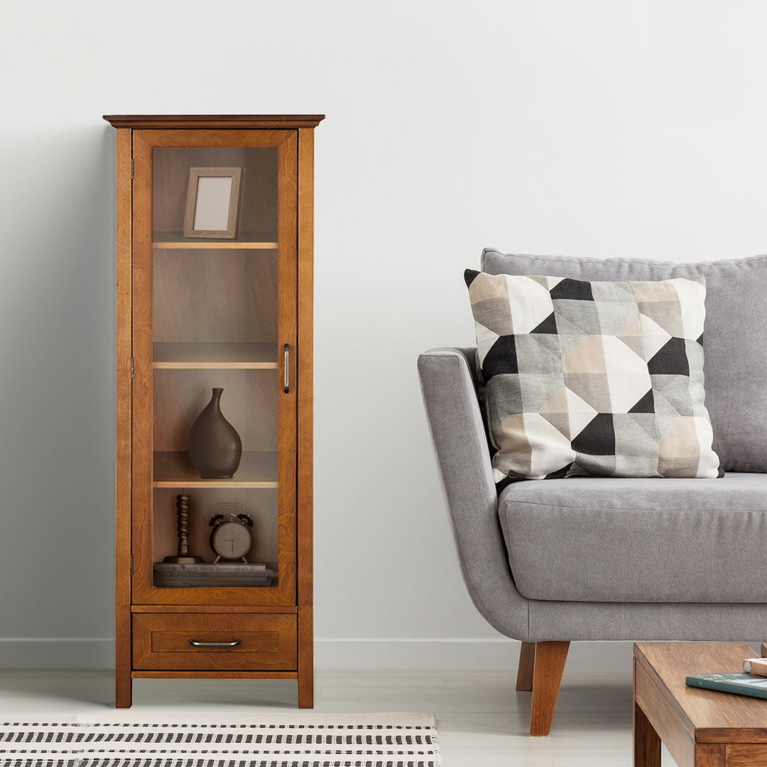 A wooden floor cabinet with a glass door and a drawer and a gray sofa with one pillow in a room.