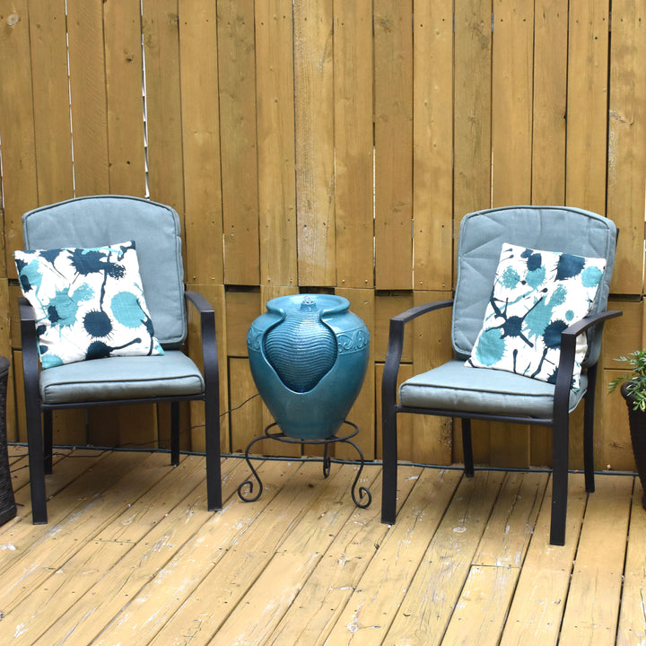 A teal vase-shaped water fountain between two chairs on a wooden patio