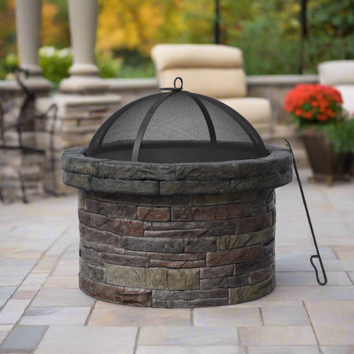 A stone fire pit with a mesh cover and metal poker on a tiled patio, with outdoor seating and potted plants in the background.