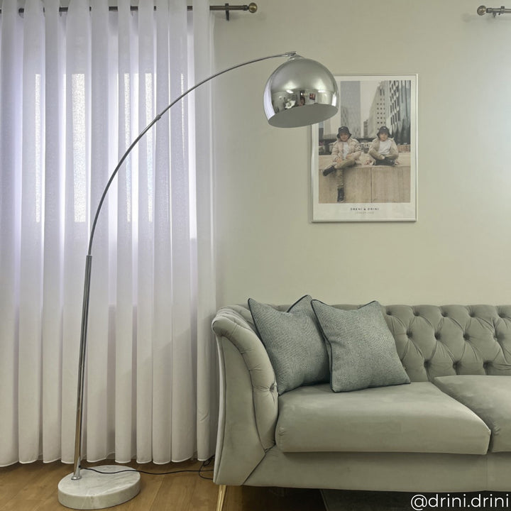 A modern living room features a tufted gray couch, a curved floor lamp with a metallic shade, and a framed wall photo. Light filters through sheer white curtains.