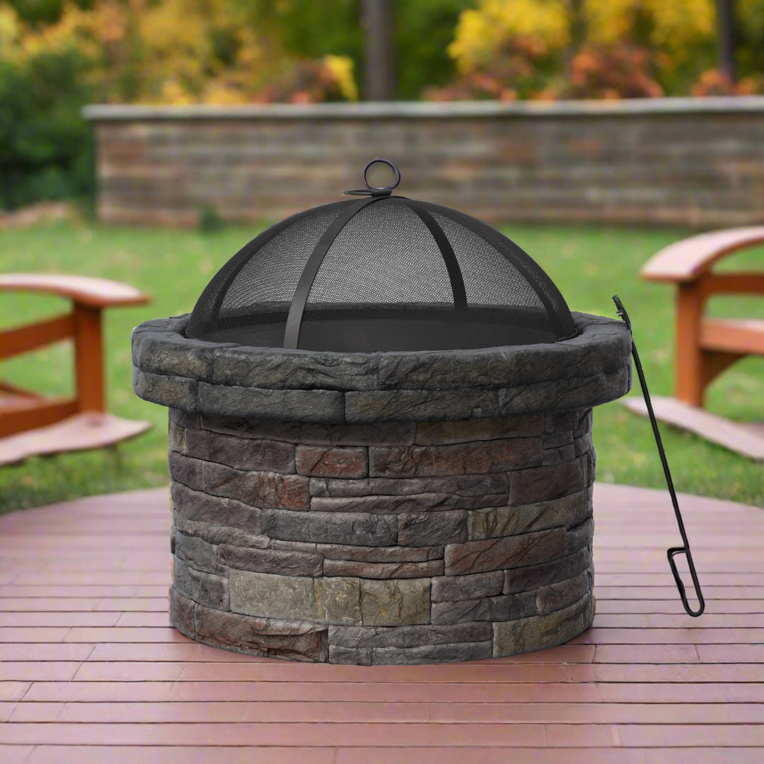 A round stone fire pit with a mesh cover is placed on a wooden deck in a backyard with green grass and trees in the background. A metal fire poker rests against the fire pit.