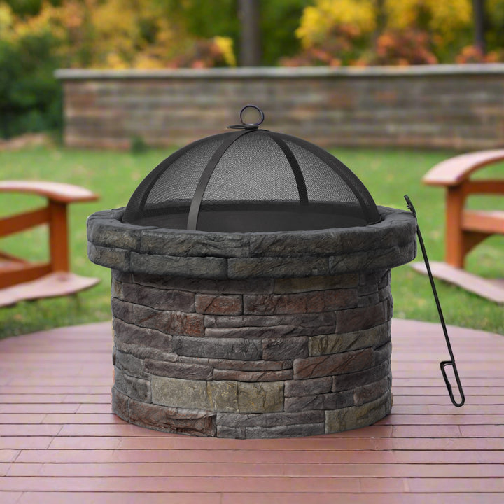 A round stone fire pit with a mesh cover is placed on a wooden deck in a backyard with green grass and trees in the background. A metal fire poker rests against the fire pit.