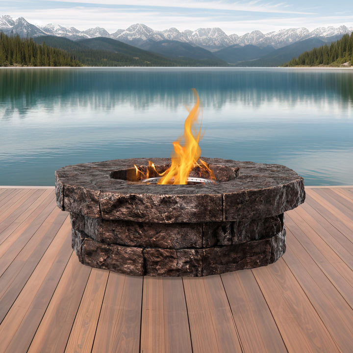 A Teamson Home Outdoor Circular Stone-Look Propane Gas Fire Pit, Red-Brown with flames on a wooden deck overlooking a tranquil lake with mountain views.