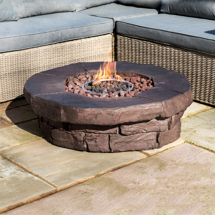 A Teamson Home Outdoor Circular Stone-Look Propane Gas Fire Pit, Red-Brown with flames centered amidst stone-textured edges on a patio.