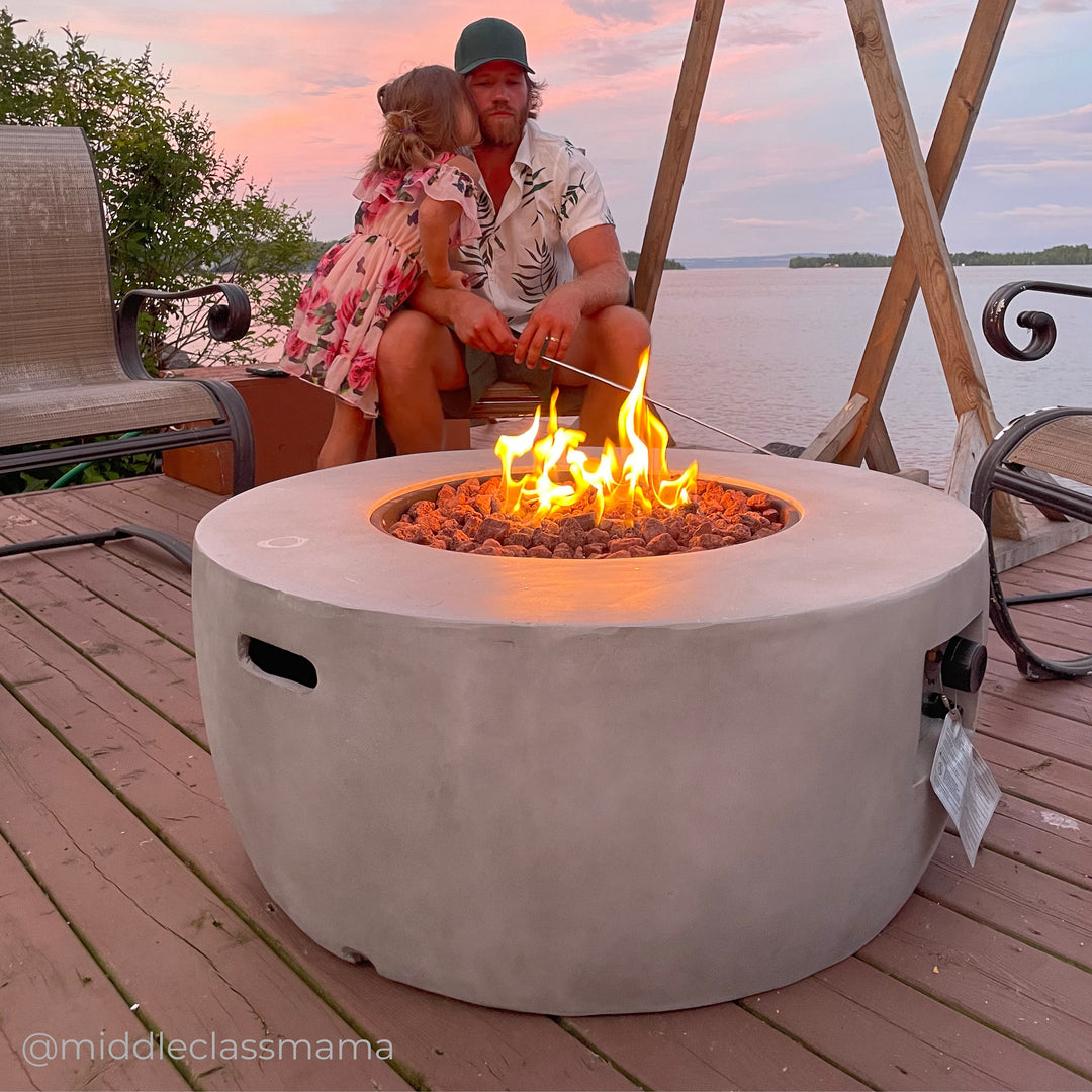 A father with a green cap and hawaiian shirt sits roasting a marshmellow with his daughter in a floral dress by his side above a gas-powered fire pit.