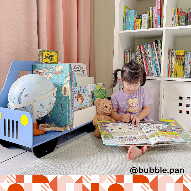 Toddler girl is reading a book on the floor. She is sitting next to a blue wooden truck bookcase that has a globe on display along with many books. 