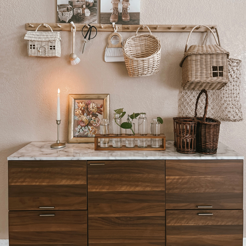 A brown side table with a faux marble top is decorated with warm brown and beige tones. The side table  has vases on top, a candle, a flower picture frame and wicker baskets. 