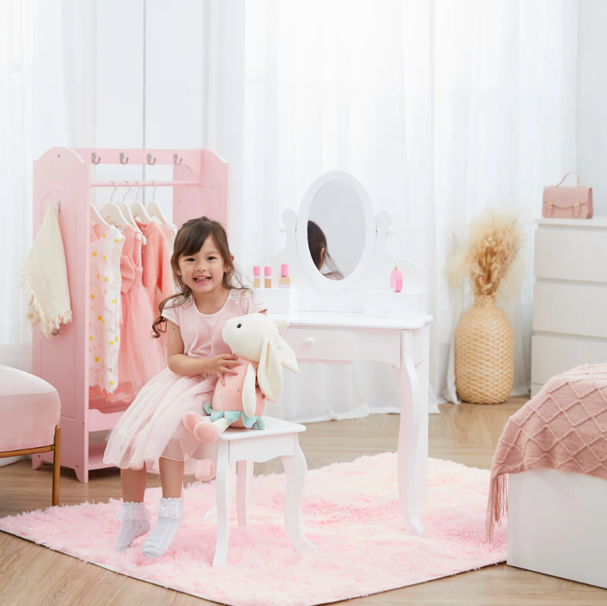 A young toddler girl is wearing a pink dress. She is sitting at a white teamson vanity and holding a white bunny rabbit toy.