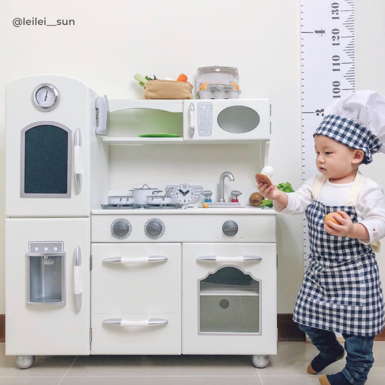 Toddler boy dressed up in a navy and white chef's hat and apron is standing next to a white kids play kitchen. 