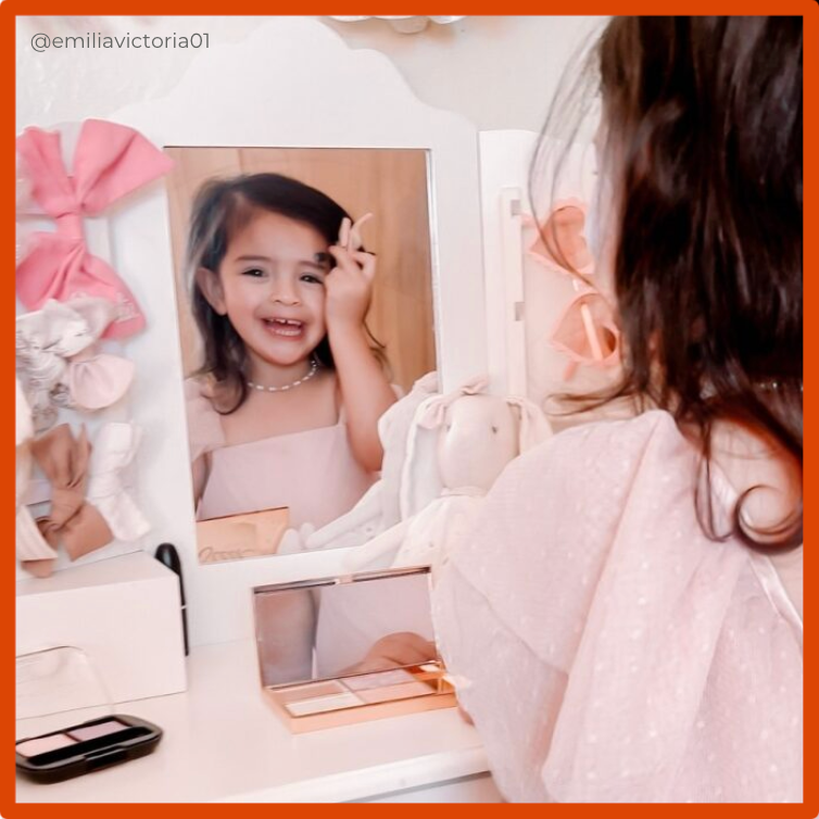 A little girl looking into a vanity mirror dressed in a pink dress clipping a pink bow in her dark hair